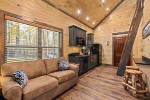 Ein Cabin-Interieur mit Holzboden, einer Couch, einer Küche mit modernen Geräten und einem Fenster mit Jalousien.