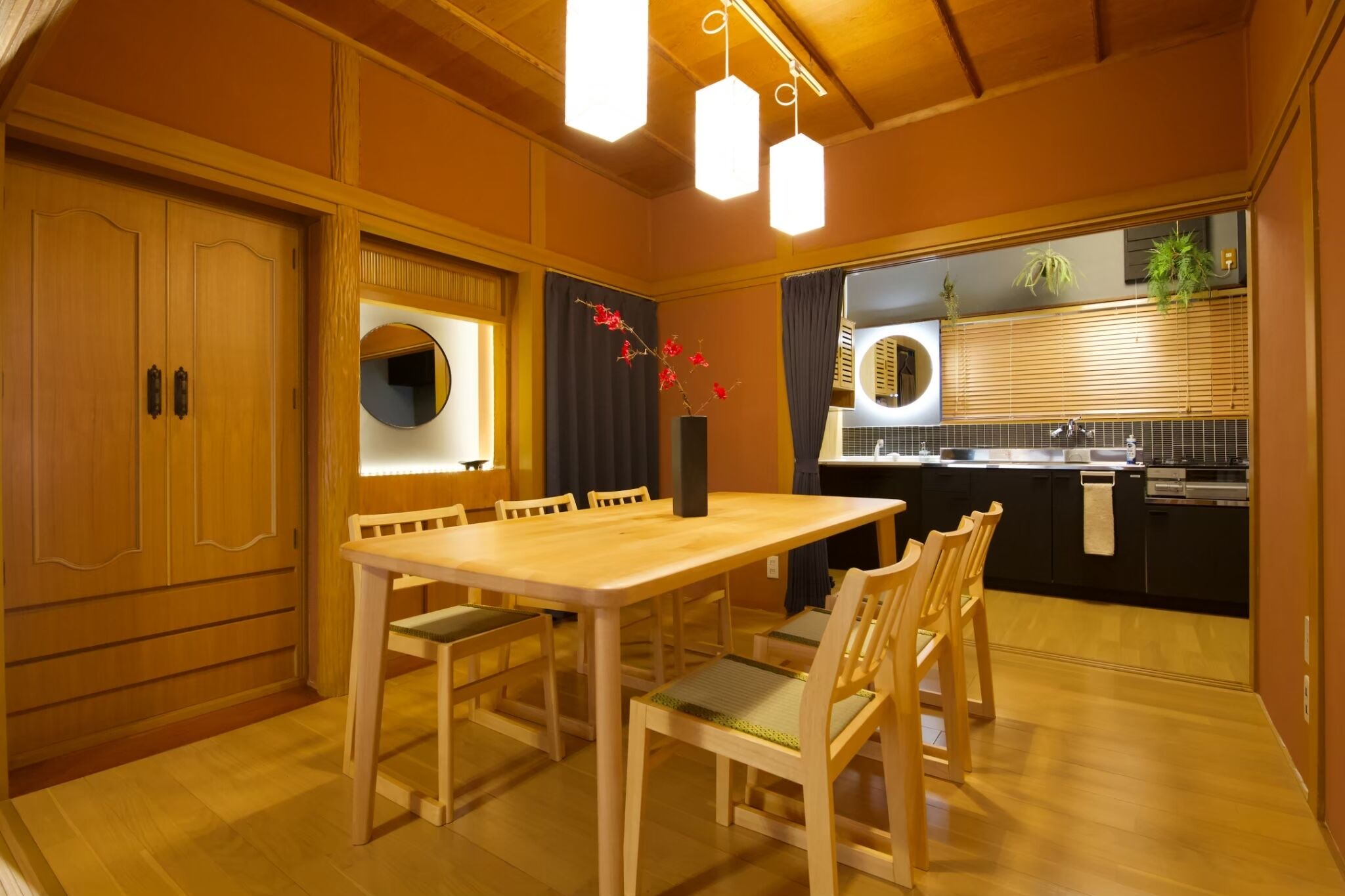 Una mesa de comedor de madera con sillas, un espejo redondo y un jarrón con flores rojas.