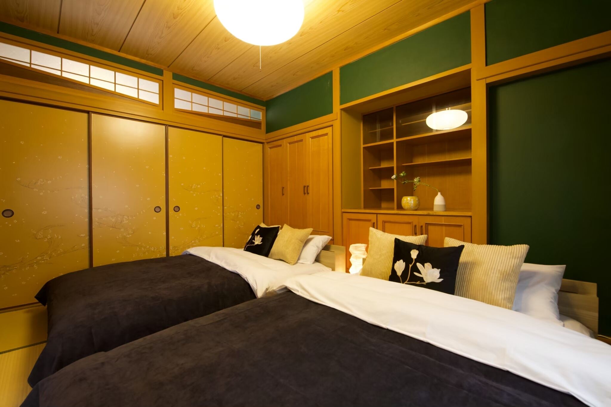 A room with two beds, wooden cabinets, a shelf with decorative items, and a green wall.