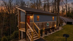 A small cabin with a wooden deck and stairs, surrounded by trees and a gravel path. - Hot Tub + King Bed | Gatlinburg 23 min (Cosby)