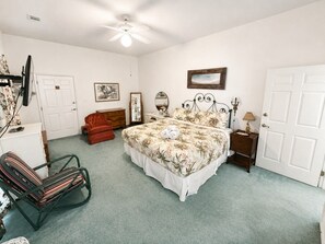Um quarto com cama, cadeira, cômoda, mesa de cabeceira e porta.