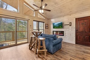 Una habitación amplia con pisos de madera, un sofá azul, una mesa de madera con taburetes, una chimenea y un televisor montado en la pared.