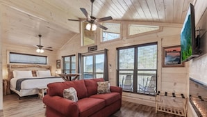 A cabin interior with a bed, red sofa, ceiling fans, large windows, and a TV. - Gatlinburg 24 min | HotTub +King Bed #8 (Cosby)