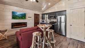 A cozy cabin interior with a fireplace, a television, a kitchen area, and a ceiling fan. - Gatlinburg 24 min | HotTub +King Bed #8 (Cosby)