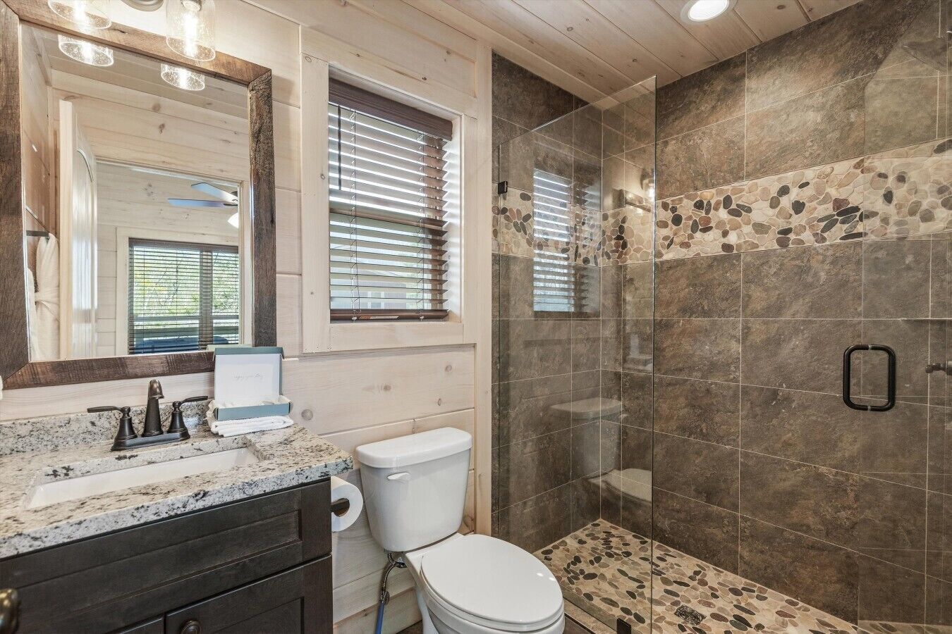 Une salle de bain dotée d’un comptoir en granit, de toilettes, d’une douche aux carreaux de pierre et d’un grand miroir.