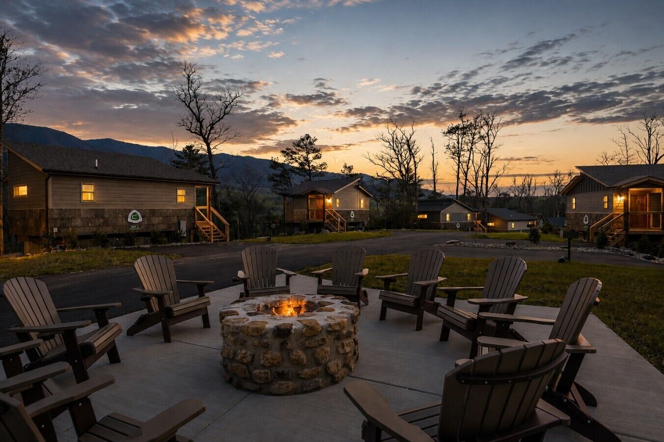 A campground with a stone fire pit surrounded by Adirondack chairs, connected to cabins with outdoor lighting.