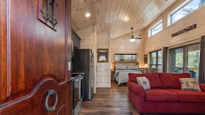 L’intérieur d’une cabine avec une porte en bois, un coin cuisine, un lit, un canapé rouge et un ventilateur de plafond.