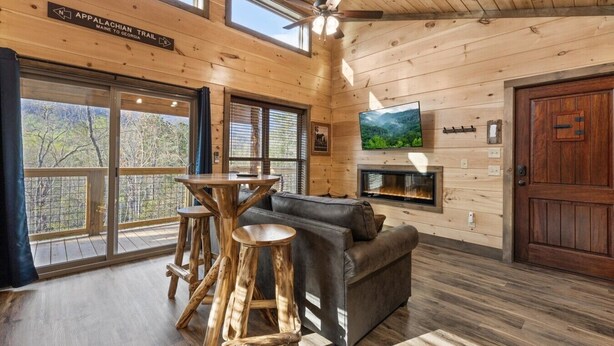 Interno di una baita con arredamento in legno, camino, televisione e vista sul paesaggio esterno.