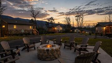 A campground with a stone fire pit surrounded by Adirondack chairs, connected to cabins with outdoor lighting.