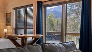 Um quarto aconchegante com decoração em madeira, uma mesa redonda, um sofá de couro e uma grande janela com vista para árvores e montanhas.