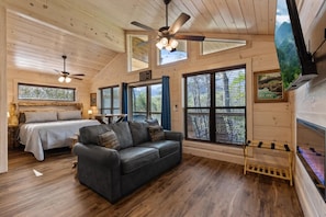 L’intérieur d’une cabane en rondins, avec un lit, un canapé, un ventilateur de plafond et de grandes fenêtres.