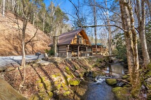 A cabin with a stone foundation, wooden exterior, and a deck overlooking a stream. - Creekside Retreat with 2 Cabins (Gatlinburg)