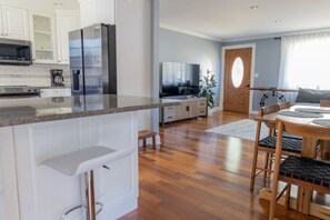 Een moderne keuken met een koelkast, magnetron en fornuis, een eethoek met een tafel en stoelen, en een woonkamer met een televisie en een plant.