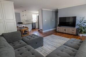 Ein modernes Wohnzimmer mit grauen Sofas, einem Flachbildfernseher und einer Küche im Hintergrund.
