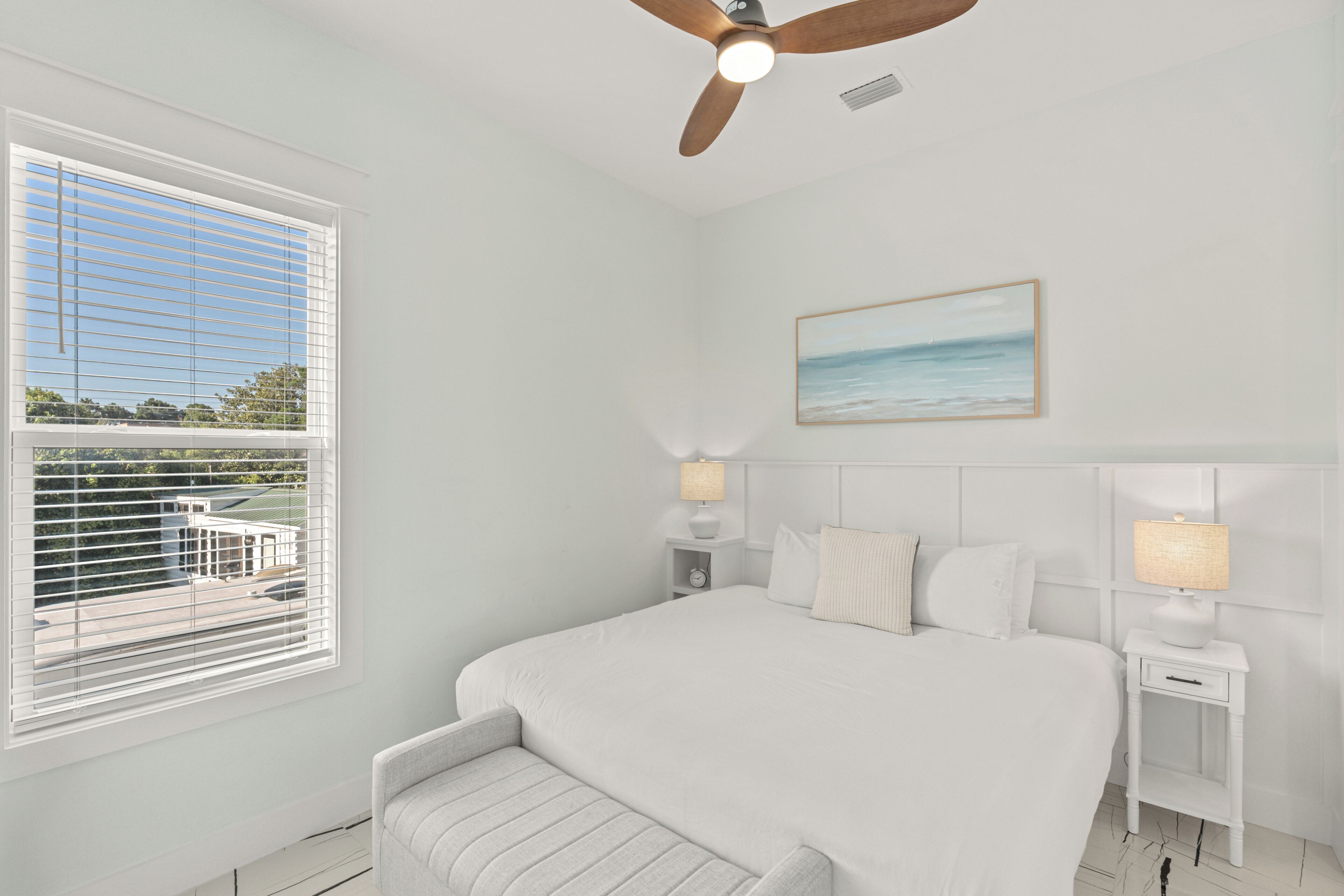 A bedroom with a bed, bedside tables, a ceiling fan, a window with blinds, and a painting on the wall.