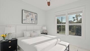 A bedroom with a bed, bedside tables, a ceiling fan, a window with a view, and a wall-mounted artwork.