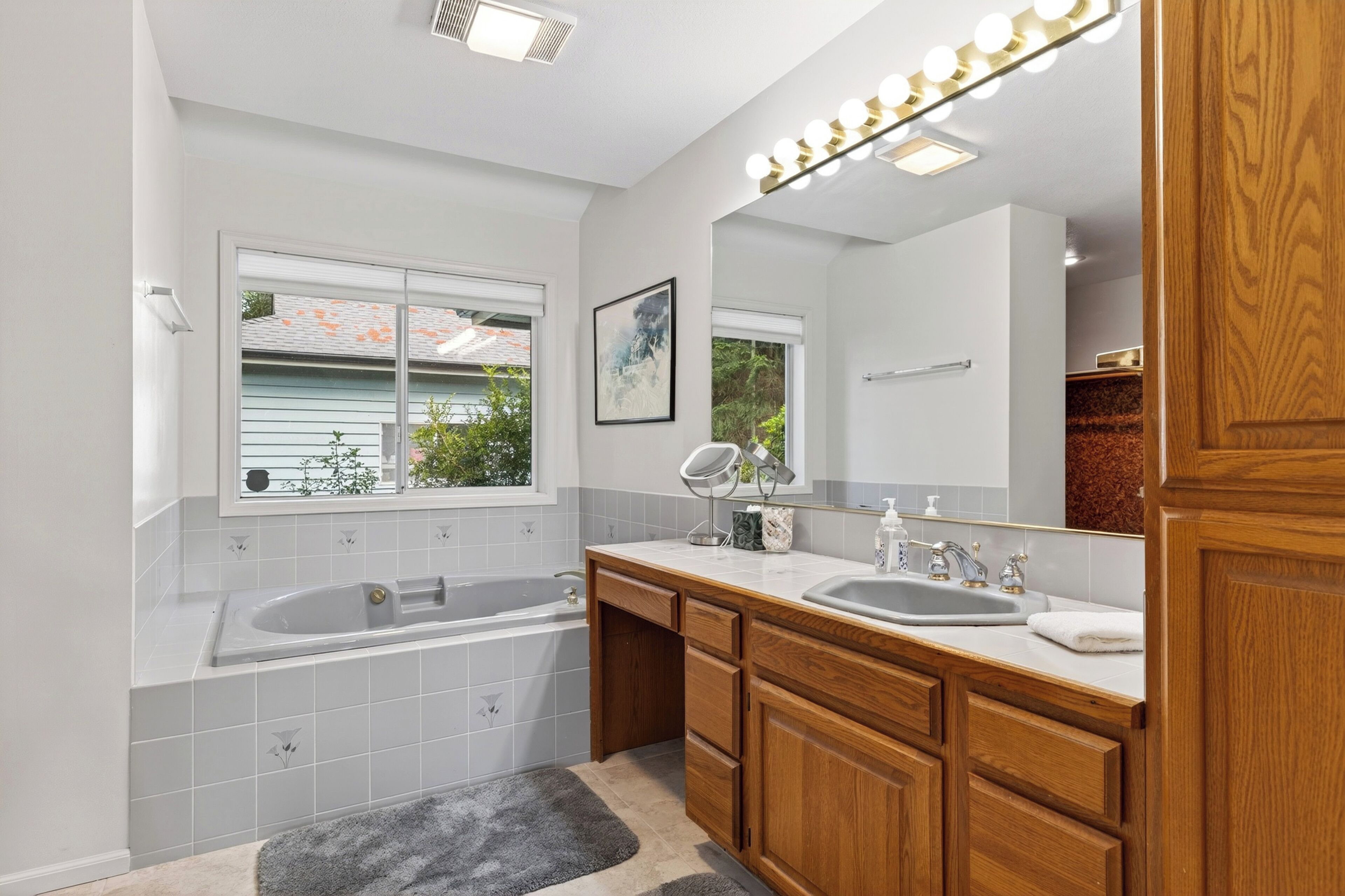 A modern bathroom with a bathtub, two sinks, wooden cabinets, a large mirror with lights, and a window with a view of the outside.