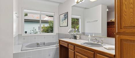 A modern bathroom with a bathtub, two sinks, wooden cabinets, a large mirror with lights, and a window with a view of the outside.