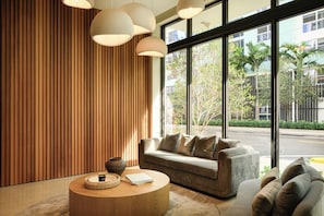 A modern interior with a sofa, a round wooden coffee table, and large windows offering a view of the street.