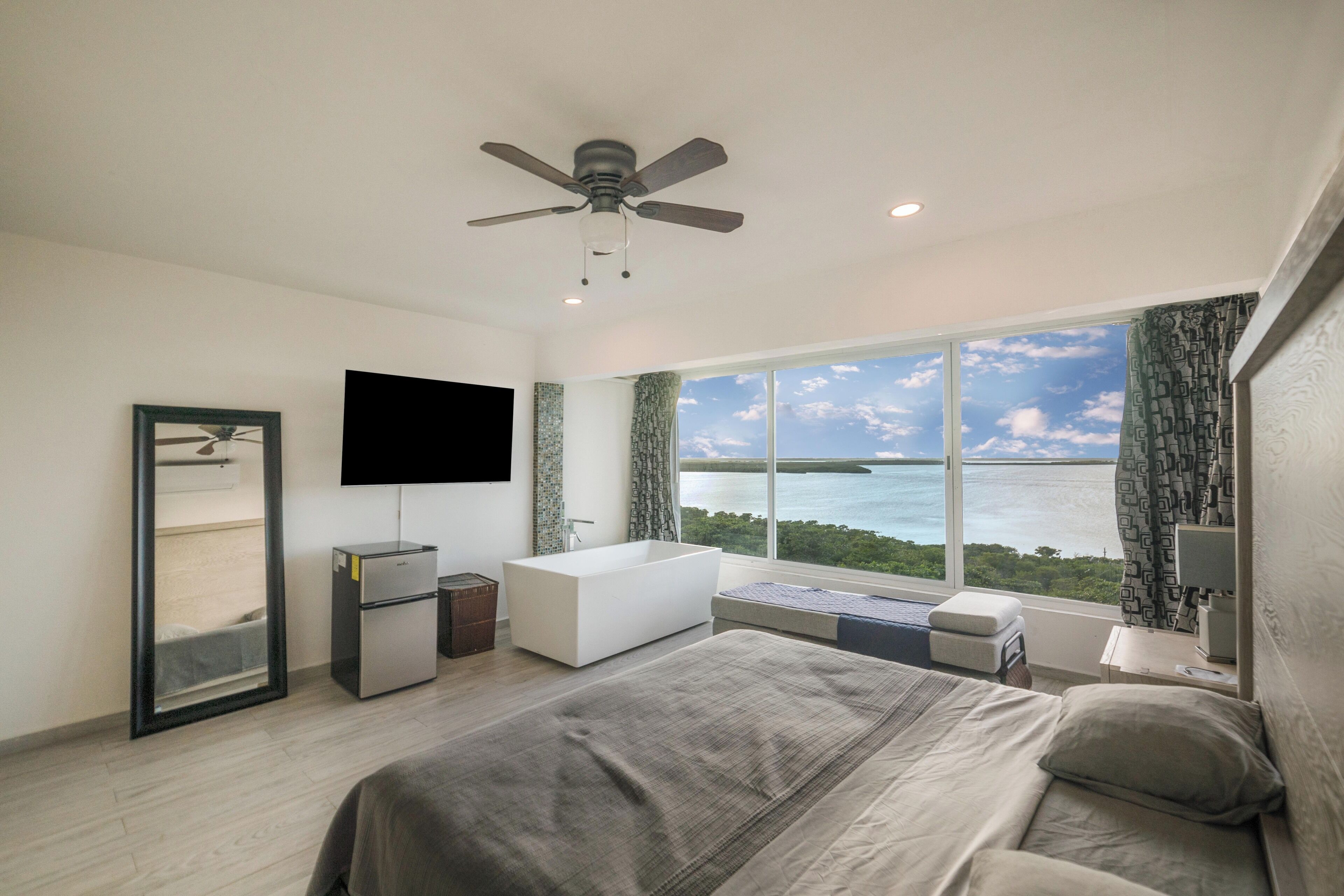 A modern hotel room with a large bed, a ceiling fan, a flat-screen TV, a built-in bathtub, and a view of the ocean.