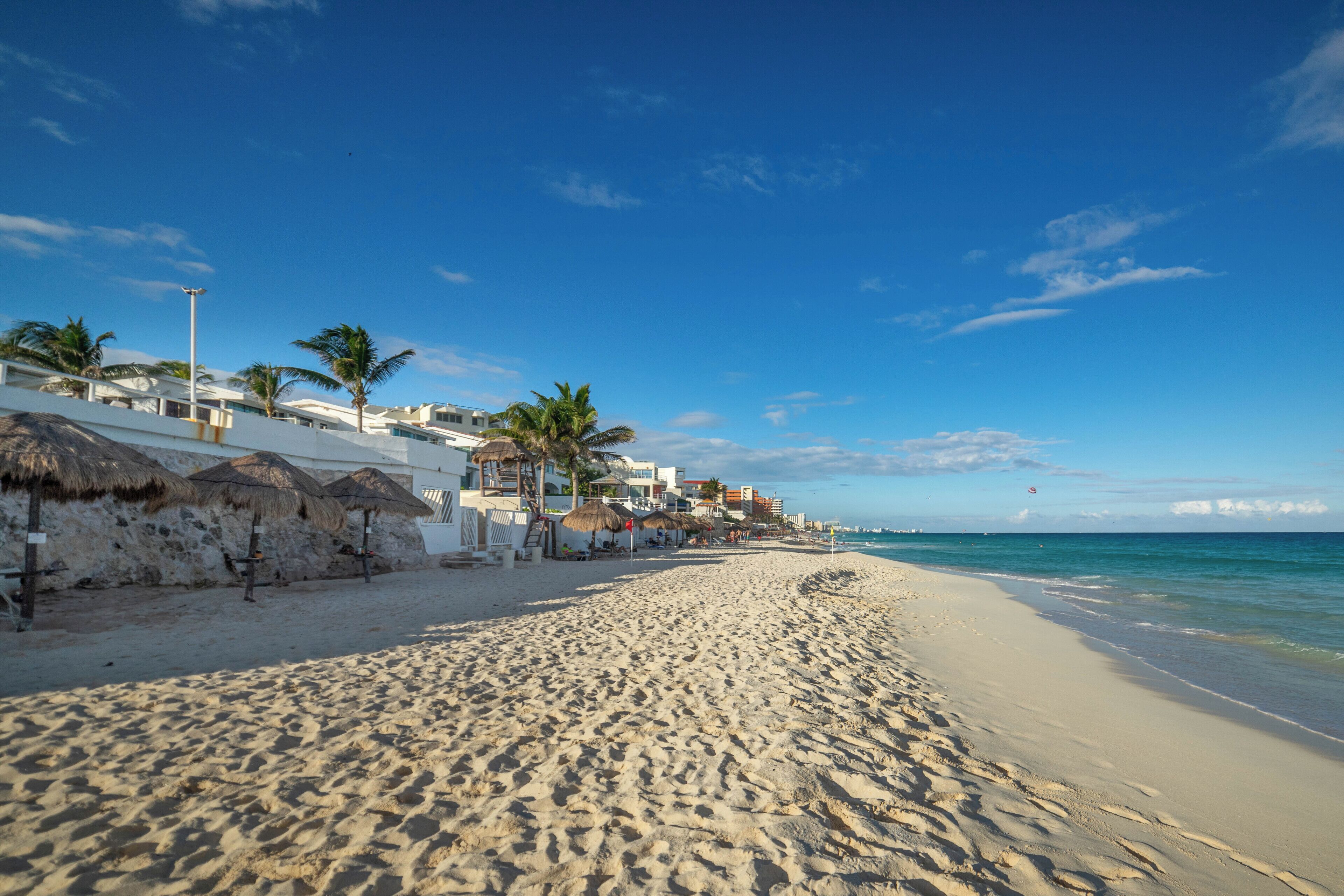 Een strand met witte gebouwen, palmbomen en rieten parasols.