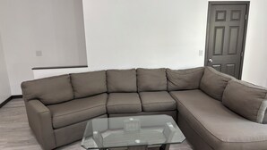 Ein Eckwohnzimmer mit einem Ecksofa, einem Glastisch und einer Tür im Hintergrund.