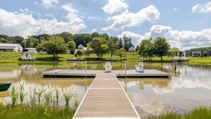 Un muelle de madera que lleva a un lago tranquilo con cielo despejado, rodeado de vegetación y casas.