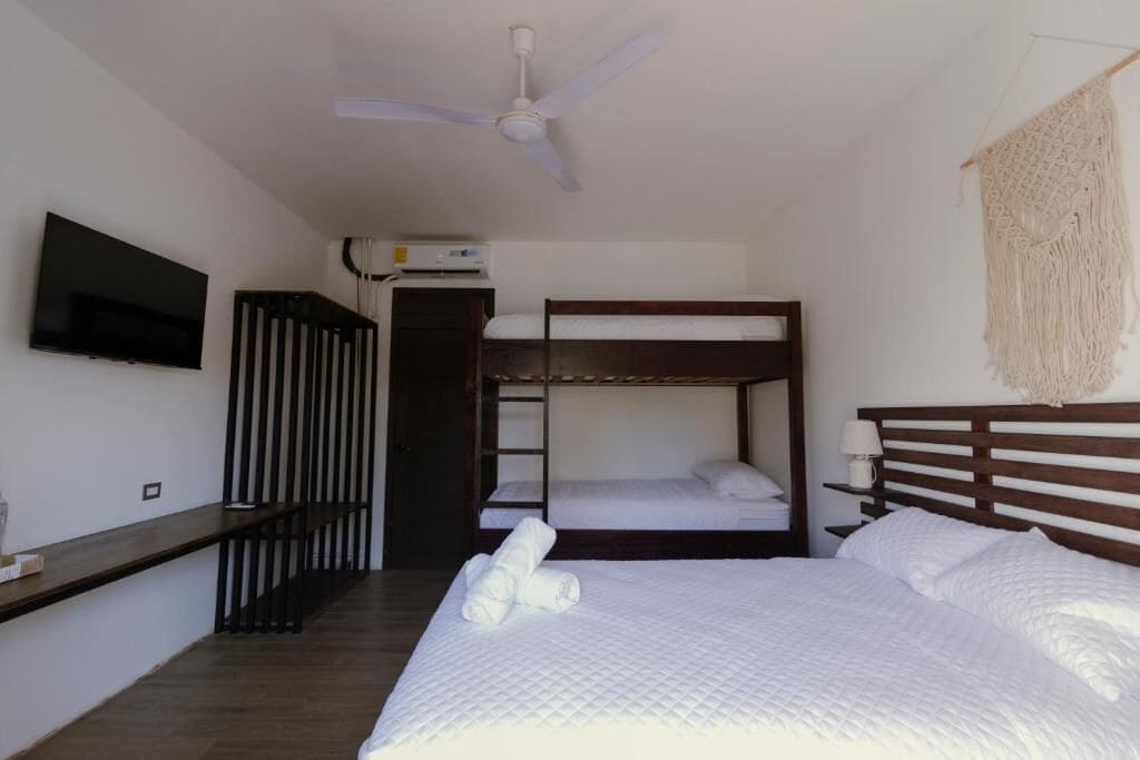 Habitación de hotel con literas, un televisor de pantalla plana, ventilador de techo y un tapiz de macramé.