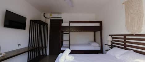 Habitación de hotel con literas, un televisor de pantalla plana, ventilador de techo y un tapiz de macramé.