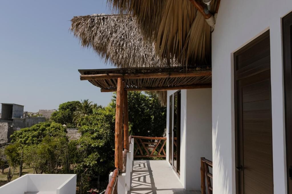 A balcony with a thatched roof, a white wall, and a view of greenery and buildings.