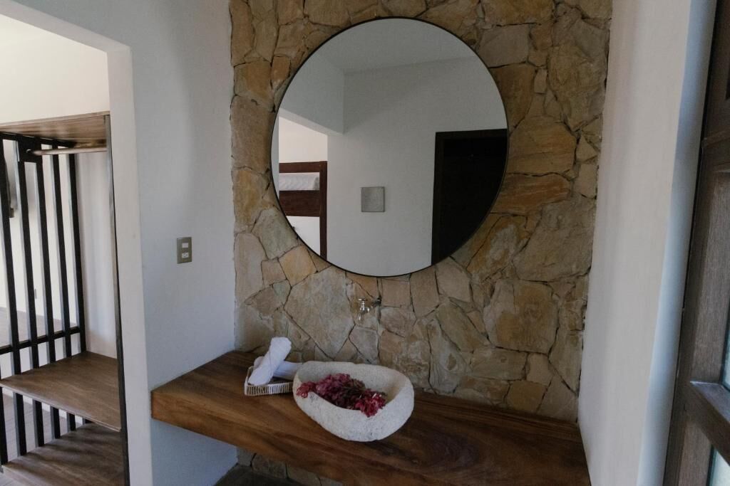 Un bany amb paret de pedra, un mirall rodó, un tocador de fusta i una bassina de pedra amb flors.
