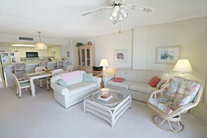 A living room with a white sofa, a wicker chair, a glass coffee table, and a dining area with a white table and chairs. - Incredible Direct Oceanfront Views from 6th Floor Balcony at Sand Dollar 3-604 (St. Augustine)