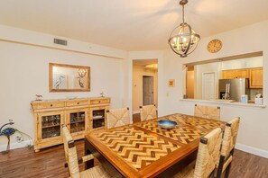 A dining room with a wooden table, woven chairs, a chandelier, a wall clock, and a kitchen visible through an open doorway. - Updated, spacious 3/2 in Gated Seaside at Anastasia A103 (St. Augustine)