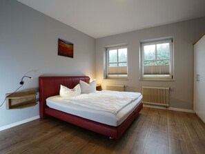 Een slaapkamer met een rood bed, twee ramen, een radiator en een wandplank.