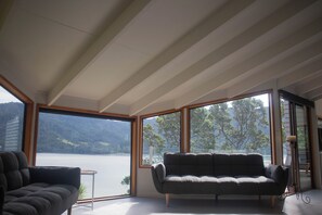 Ein modernes Wohnzimmer mit einer Couch, einem kleinen Tisch und großen Fenstern mit Blick auf einen See und Berge.