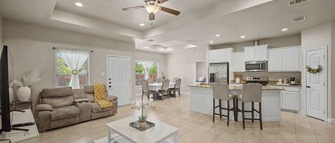A spacious living area with a sofa, coffee table, and TV, a dining area with chairs and a table, and a kitchen with white cabinets and appliances.
