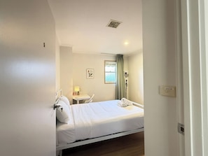Ein Hotelzimmer mit Bett, Nachttisch, Lampe, Fenster mit Vorhängen und einem kleinen Tisch mit Lampe.