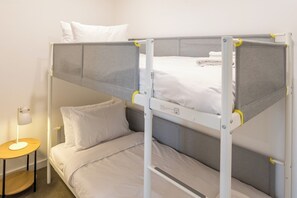 Kerrossänky, jossa on pääty, yöpöytä lampun kanssa ja seinälle kiinnitetty hylly.