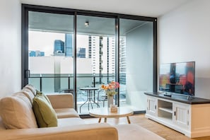Een moderne woonkamer met een bank, een salontafel met bloemen, een televisie op een kast en een stadszicht door grote glazen deuren.