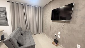A modern living room with a grey sofa, a flat-screen TV mounted on the wall, and a window with curtains.
