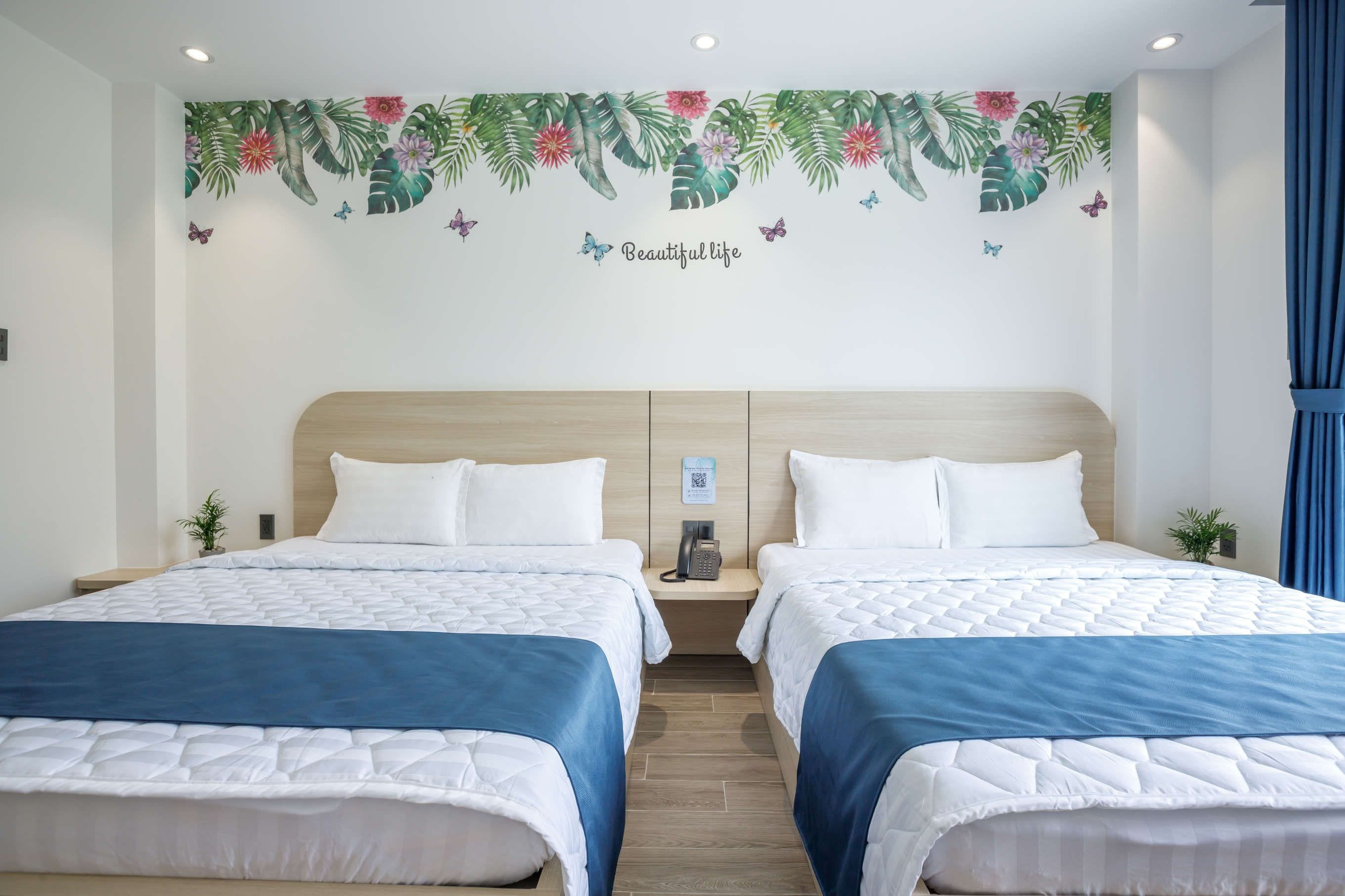 A hotel room with two beds, a wooden headboard, a wall with a tropical floral mural, and a blue curtain.
