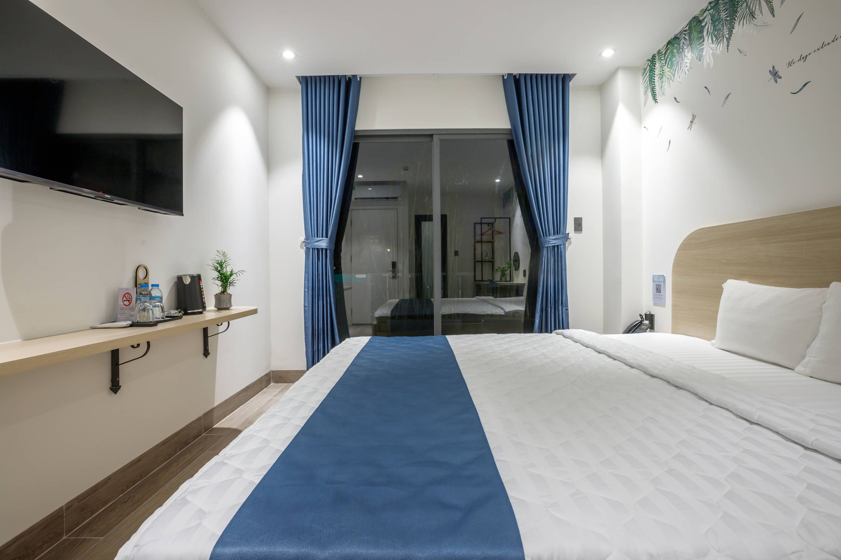 A hotel room with a bed, a TV mounted on the wall, a bedside table with a kettle and a plant, and blue curtains.