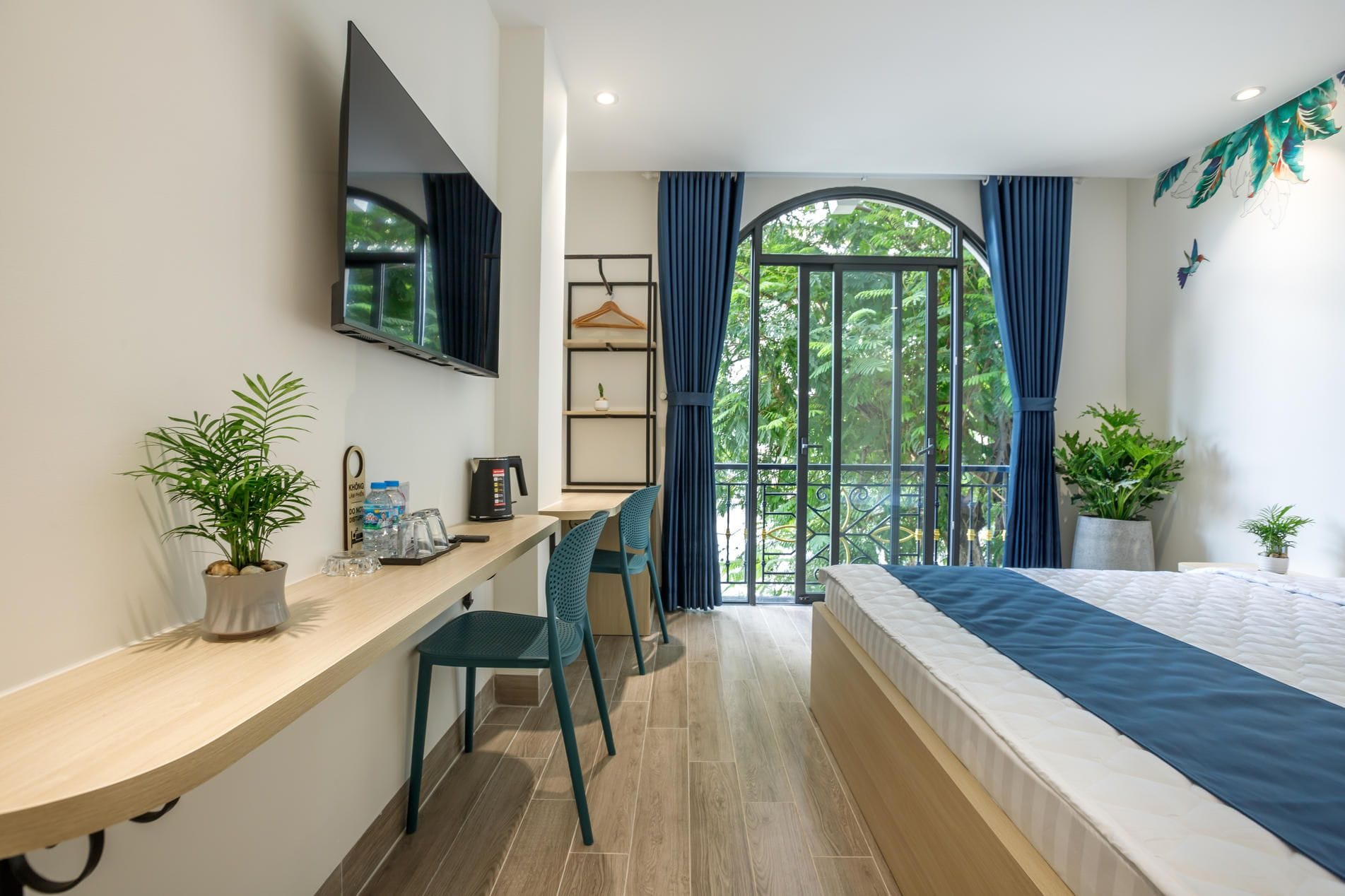Habitación de hotel con cama, escritorio con silla, televisor y balcón con vista a áreas verdes.