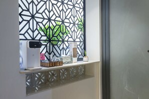 Ein Zimmer mit Fenster, einer weißen Wand, einem schwarzen, geometrisch gemusterten Bildschirm, einem weißen Regal mit diversen Gegenständen und einer Tür.