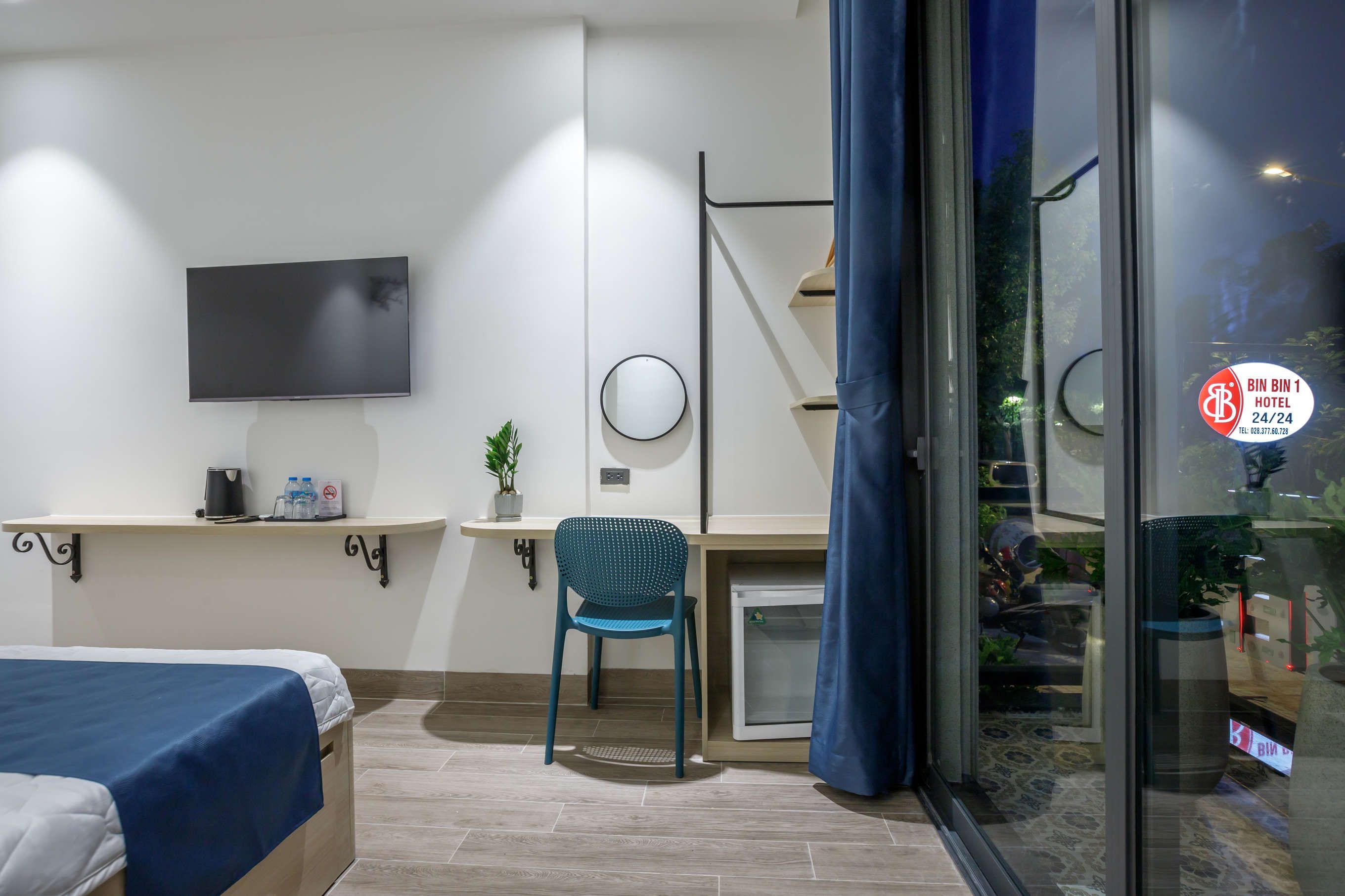 Hotellihuone, jossa on sänky, työpöytä tuolilla, televisio, pieni pöytä, jossa on kasvi, ja liukuovi, joka johtaa parvekkeelle.