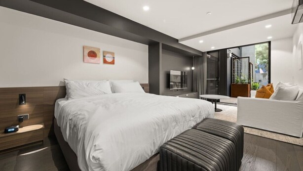 A modern hotel room with a large bed, bedside table, and a seating area with a sofa and armchair. - Terrace Studio Retreat w/ BBQ & AC (Braddon)