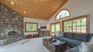 Una sala de estar con chimenea de piedra, ventanas amplias, techo de madera y sofá gris.