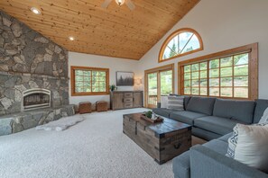 A living room with a stone fireplace, large windows, a wooden ceiling, and a gray sofa. - 4BR Tahoe Donner retreat with fireplace, deck, HOA access- Chatterjee (Truckee)