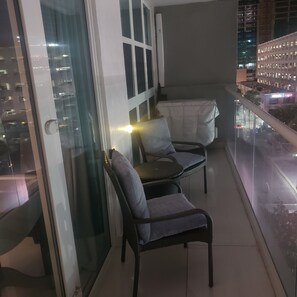 Ein Balkon mit zwei Stühlen, einem kleinen Tisch, einer Glastür und Blick auf nächtliche Stadtansichten.