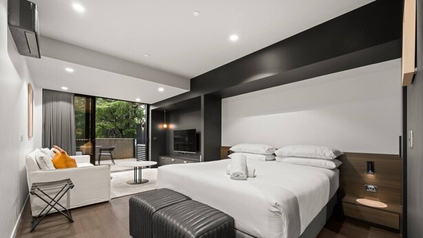 Une chambre d’hôtel moderne dotée d’un grand lit, d’un coin salon avec une table, d’une télévision et offrant une vue sur la verdure environnante.